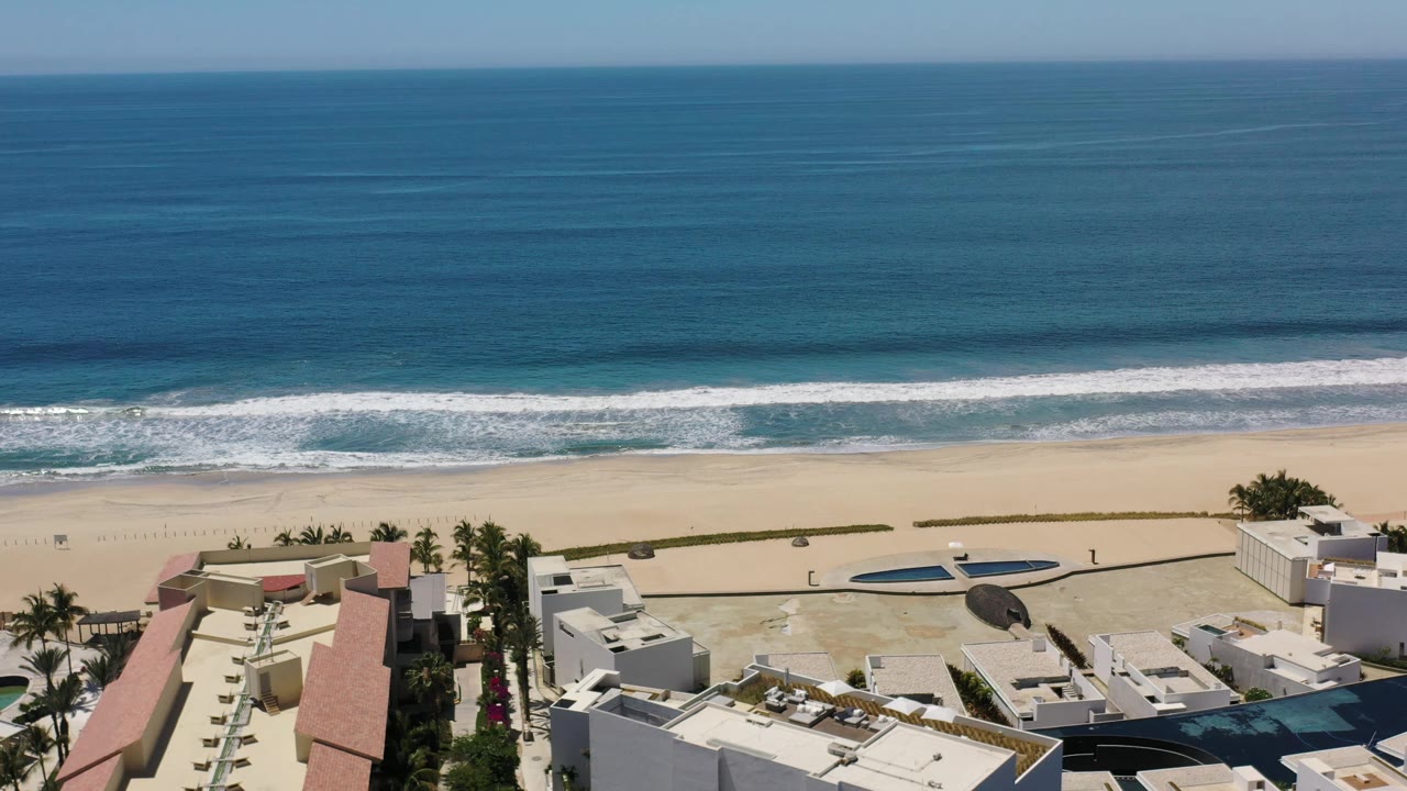 Los Cabos aerial frame showing a broad teal sea, a white surf line, open sand, palm trees, low beachfront buildings, and no visible people.