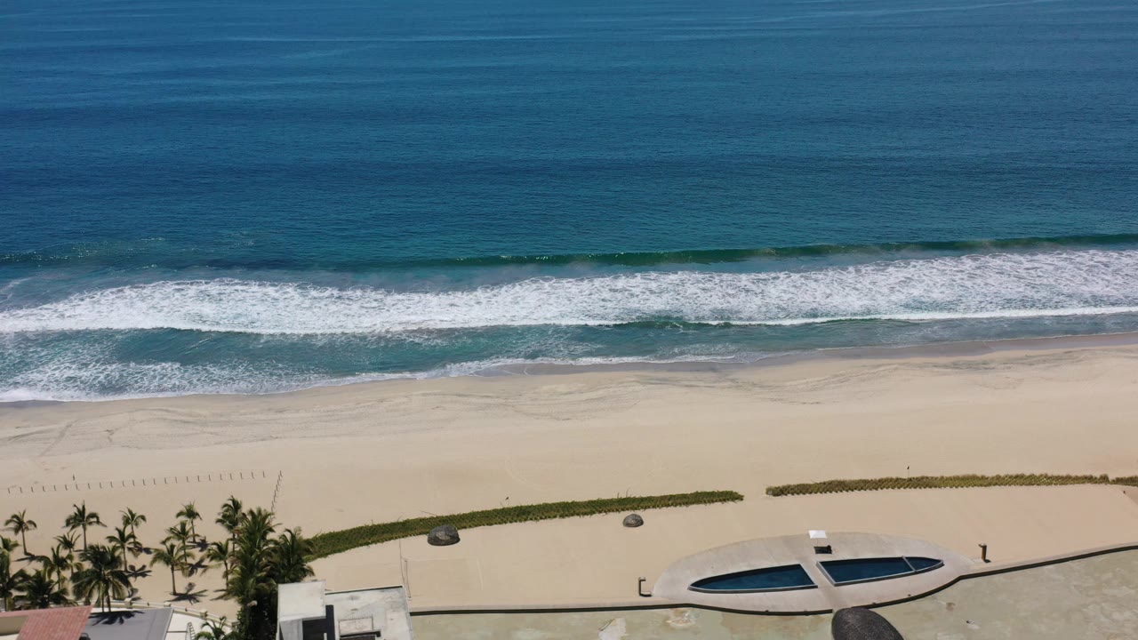 Aerial Los Cabos view where the white surf spreads wider across the shore, with blue water above, open sand below, palms and a dark oval pool shape near the lower edge, and no visible people.