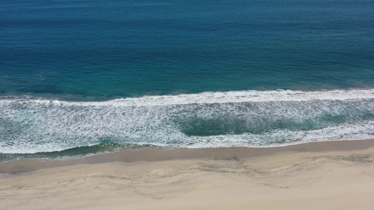 Aerial view in Los Cabos, Mexico, with deep blue water, a white surf band, beige sand, and no visible people at the start of the clip.