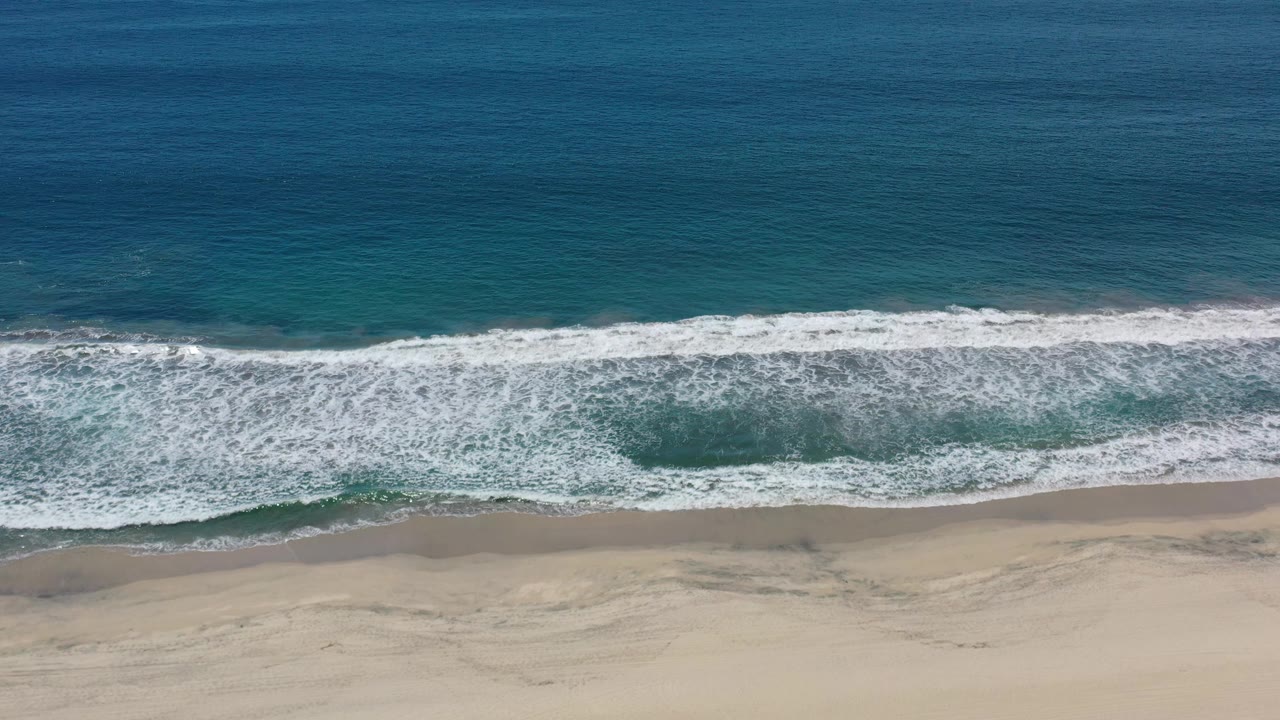 Elevated Los Cabos beach view showing a broader white wave line rolling toward pale sand, with no visible people or structures.