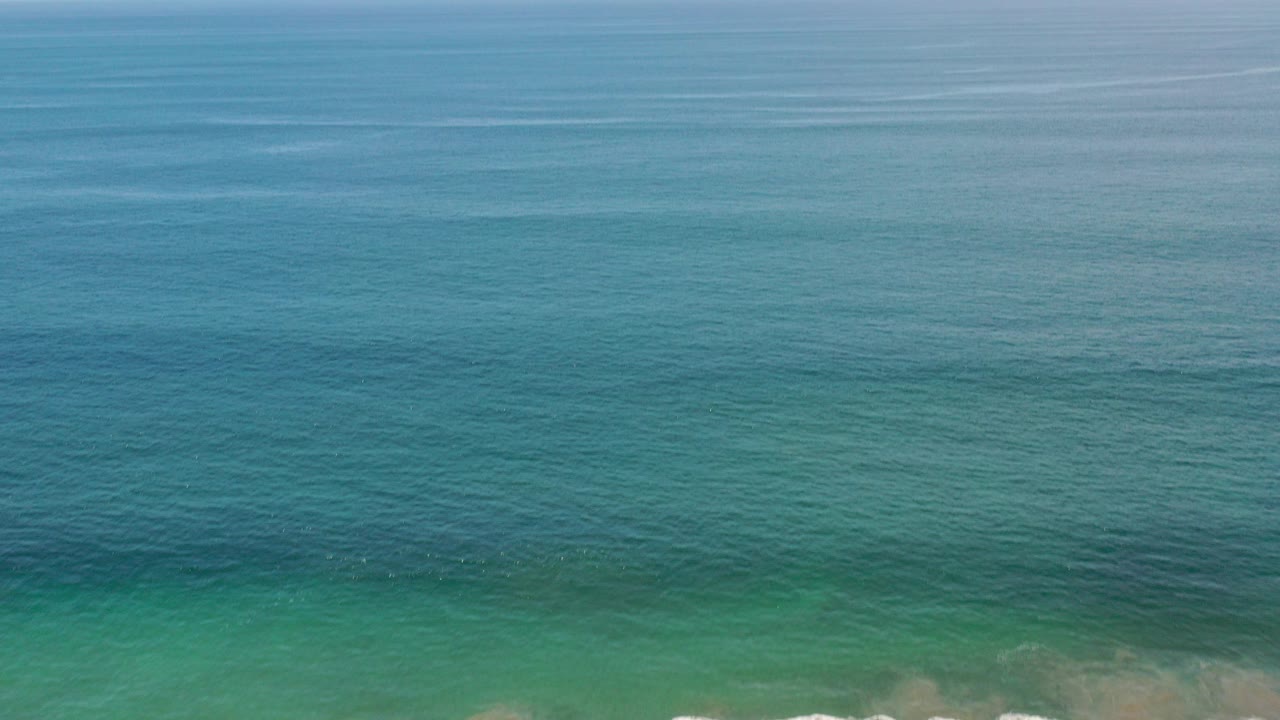 Aerial ocean view with blue-green water and a thin strip of surf at the bottom edge.