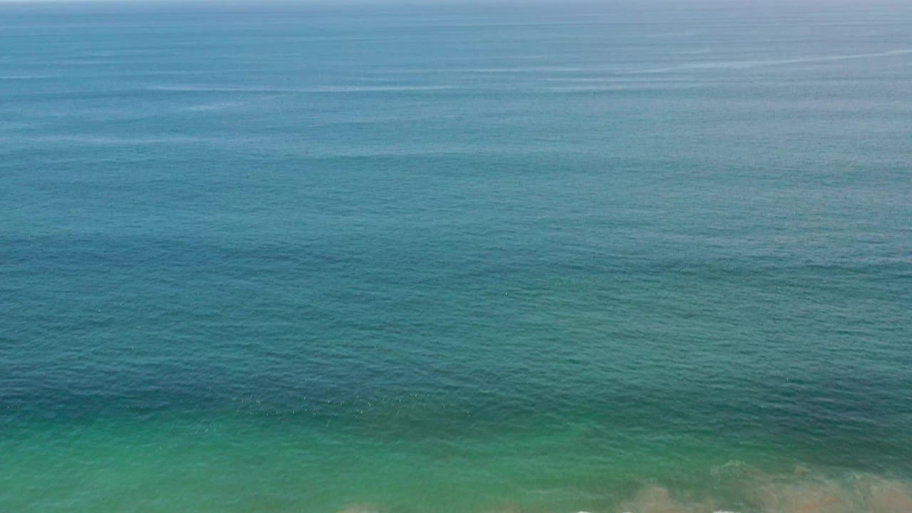 Wide aerial view of the Los Cabos Mexico ocean, with textured blue-green water, a straight horizon, pale sky, and no visible people.