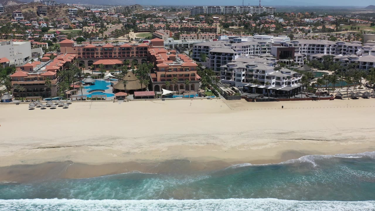 High aerial view in Los Cabos, Mexico showing resort buildings behind a wide pale beach, turquoise water in the foreground, and no clearly identifiable people.