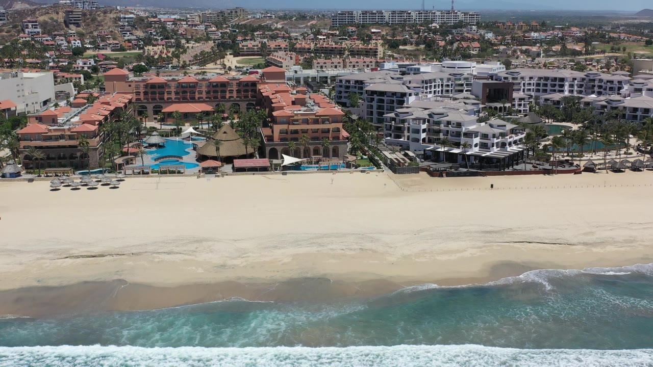 Aerial Los Cabos resort view with foamy surf crossing the lower edge, broad sand in the middle, hotel buildings and pools behind it, and no clearly identifiable people.