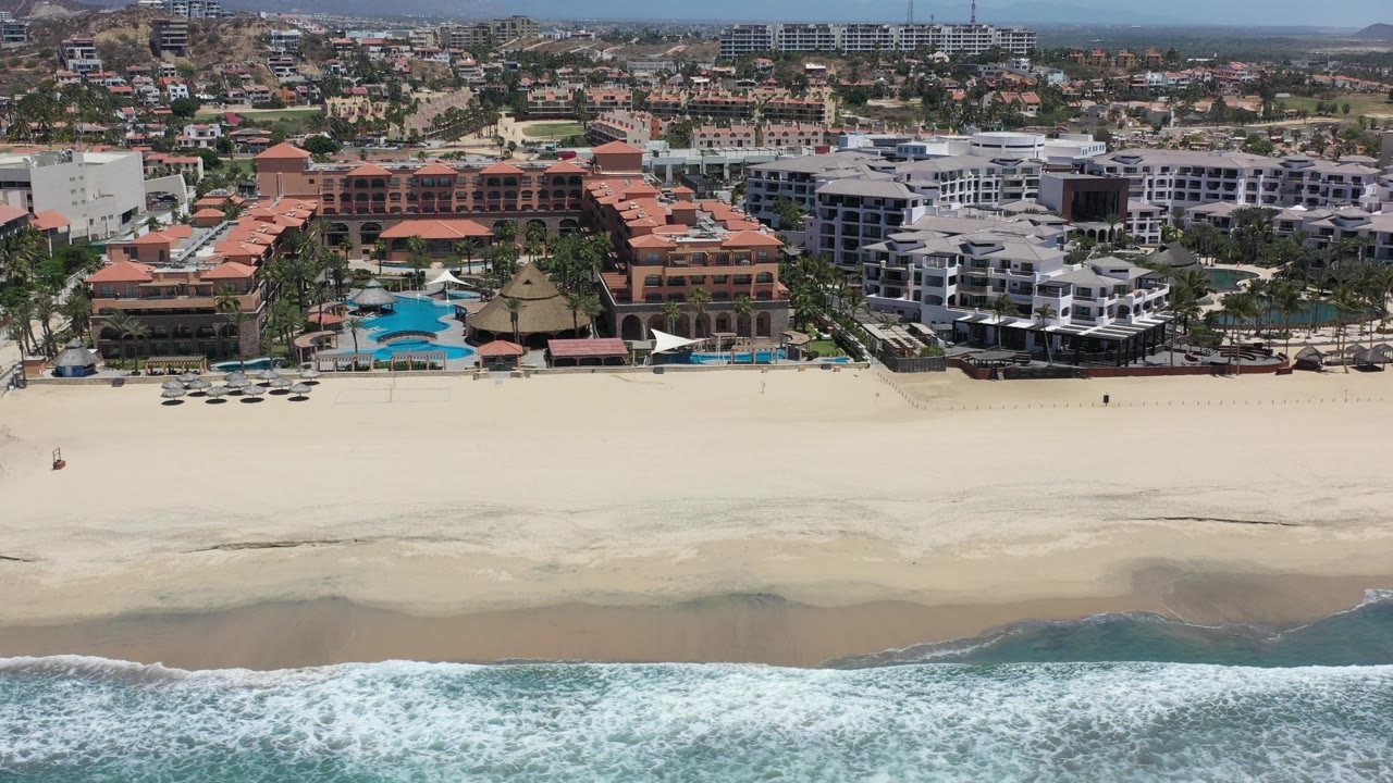 Aerial frame over Los Cabos, Mexico showing white surf in the foreground, a wide pale beach, resort buildings with red roofs, and hazy hills beyond, with no visible people.