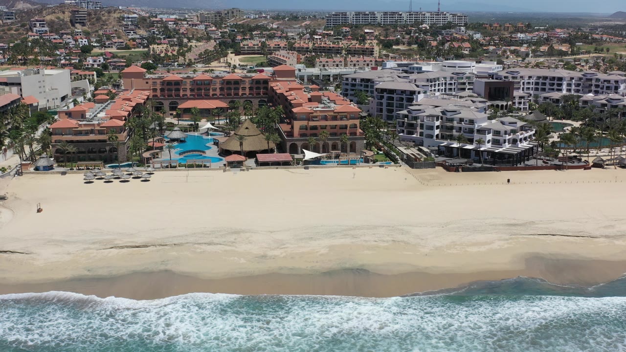 Aerial view across Los Cabos beach with a broad band of surf at the bottom, empty-looking sand, a row of beachfront hotels, and low city buildings receding toward the hills, with no visible people.