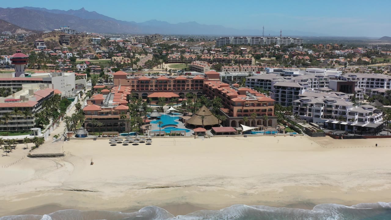 Aerial view of beachfront resort buildings, pools, sand, and mountains.