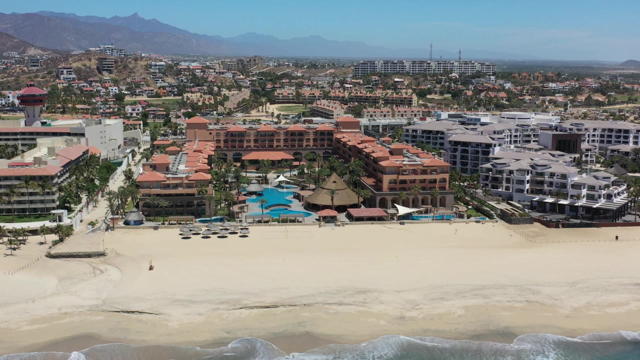 Aerial frame in Los Cabos, Mexico, with the resort pool area centered behind pale sand, low surf at the bottom edge, hillside development, and faint mountains in the distance.