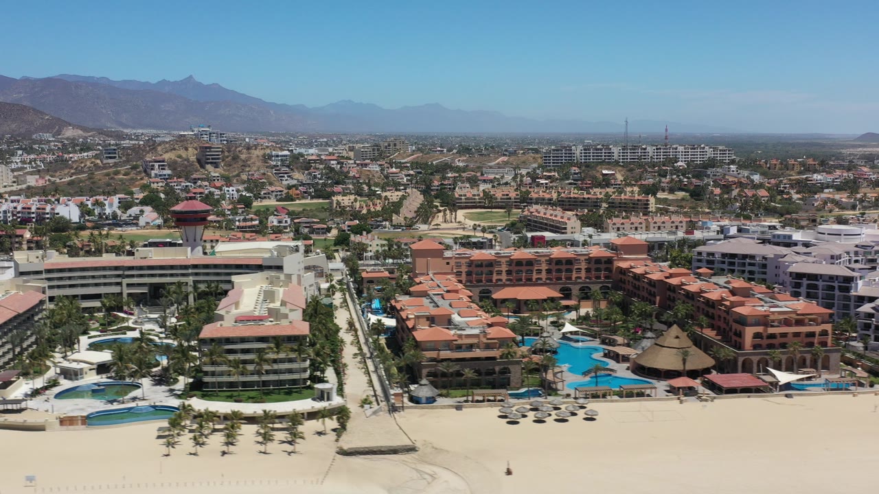 Wide aerial frame in Los Cabos, Mexico showing sandy beach, palm rows, resort pools, white and terracotta hotel buildings, inland development, hazy mountains, and no visible people.