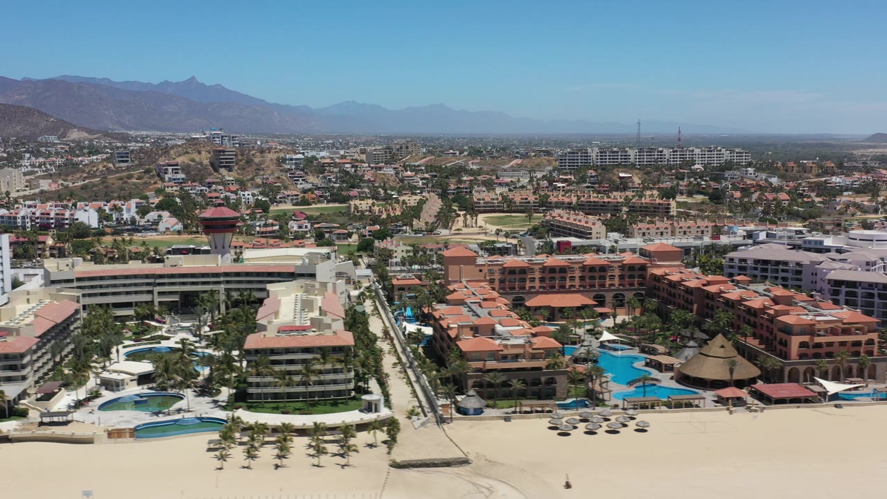 Beach resort aerial with pools, palms, hotels, city blocks, and mountains