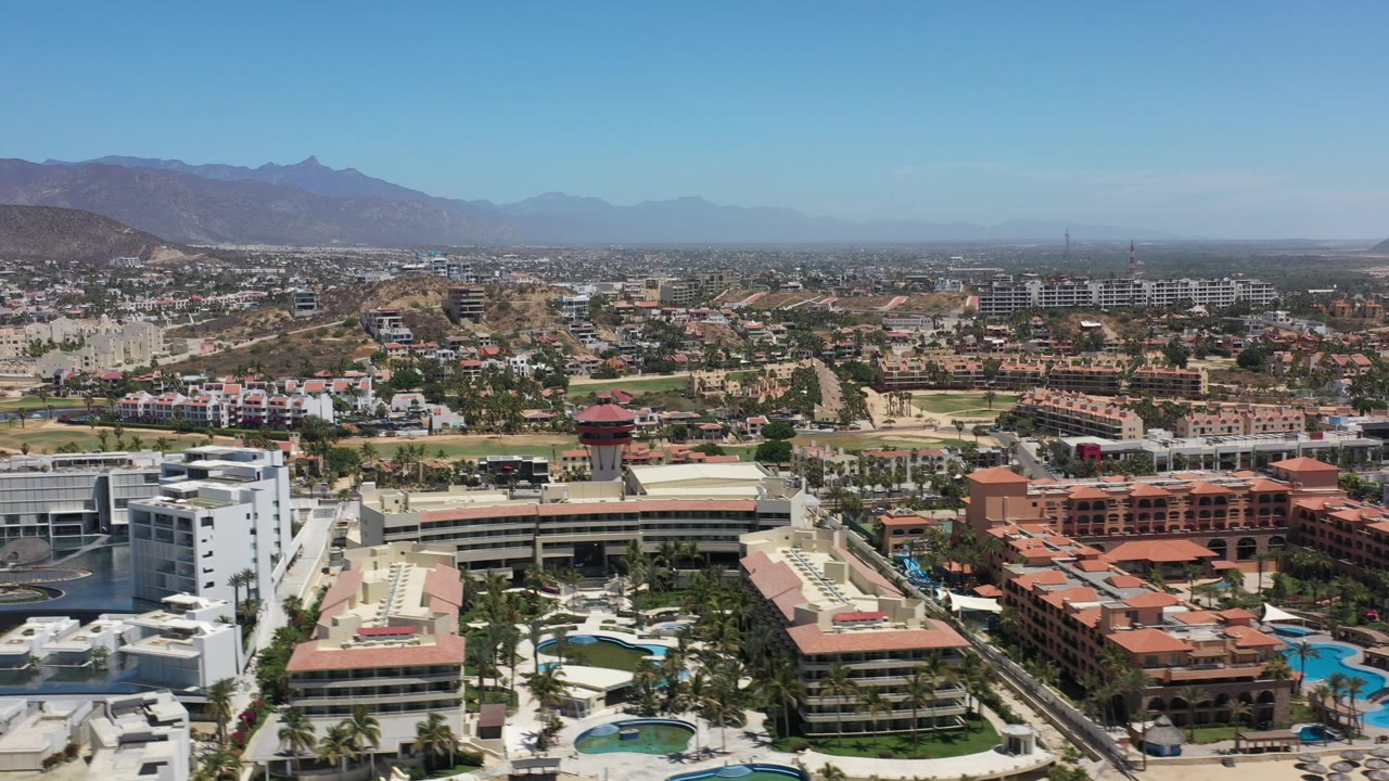 Opening aerial frame over Los Cabos, Mexico with white and terracotta resort buildings in the foreground, town blocks and roads beyond, mountains on the horizon, and no visible people.
