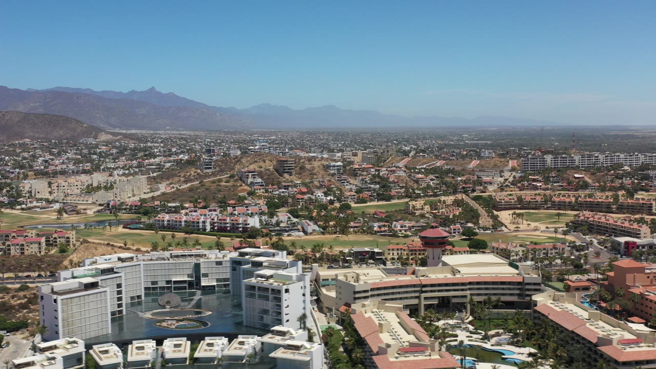 Wide aerial over Los Cabos buildings, roads, hills, and distant mountains.