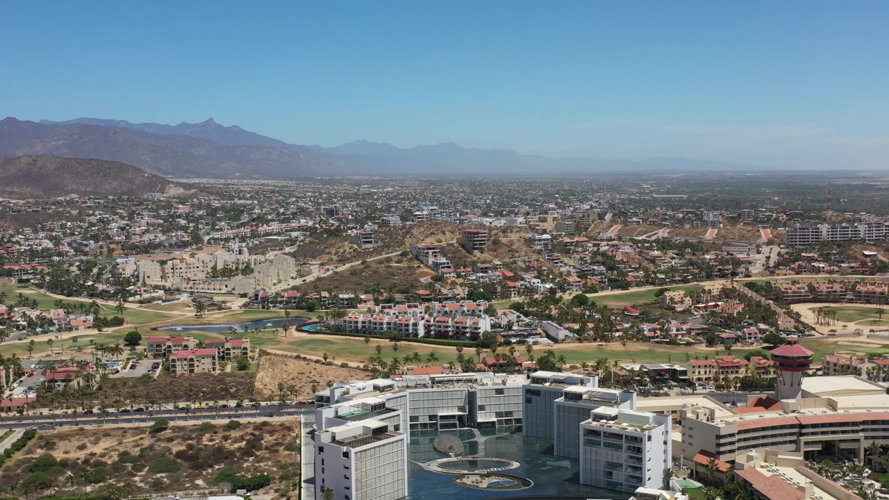 Los Cabos cityscape seen from a high vantage, with resort-like buildings at the bottom edge, roads through the midground, mountain ridges beyond, and no visible people.