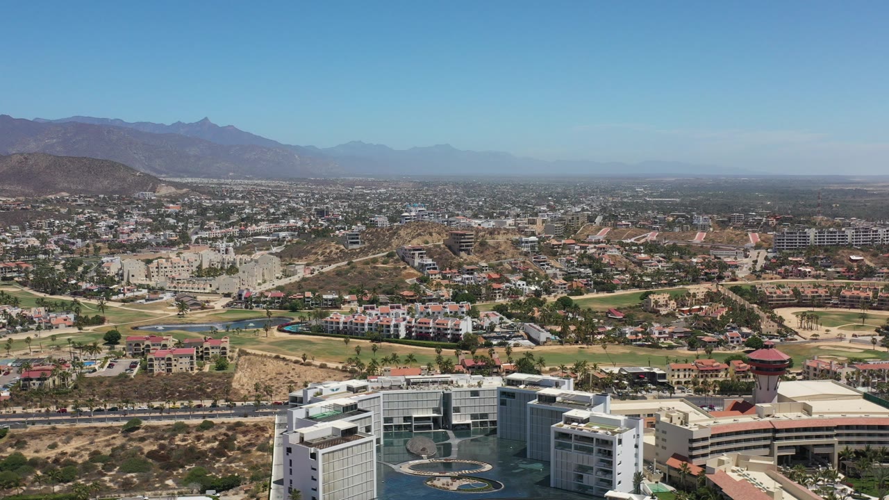 Opening aerial frame over Los Cabos, Mexico, showing a white modern building at the bottom edge, dense city blocks, tan hills, hazy mountains, clear blue sky, and no visible people.