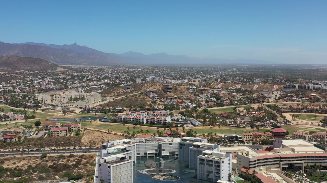 Aerial cityscape with low-rise buildings, tan hills, and a mountain horizon.