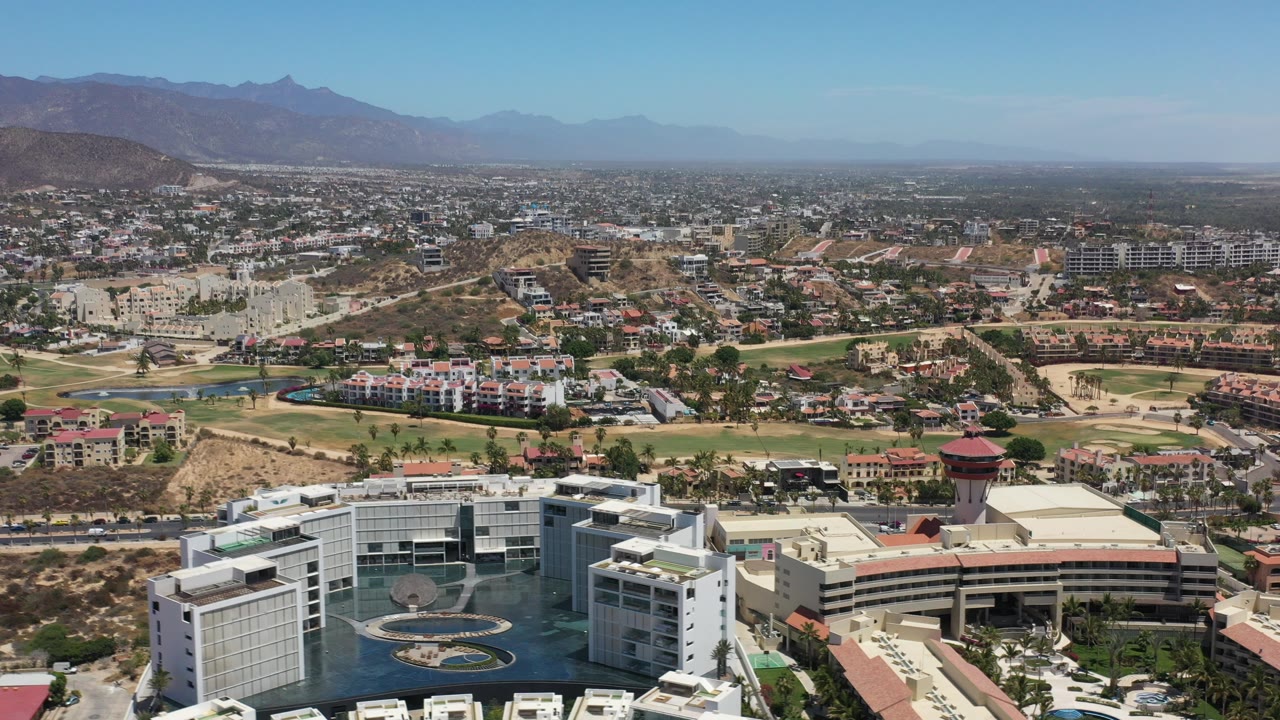 Wide aerial view in Los Cabos, Mexico, with white resort buildings and pool areas in the foreground, town development and dry hills beyond, and no visible people.