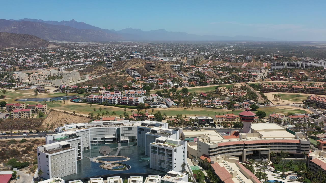 Los Cabos aerial frame centered on resort rooftops and a circular pool feature, with residential blocks, roads, tan hills, and hazy mountains in the background, no visible people.