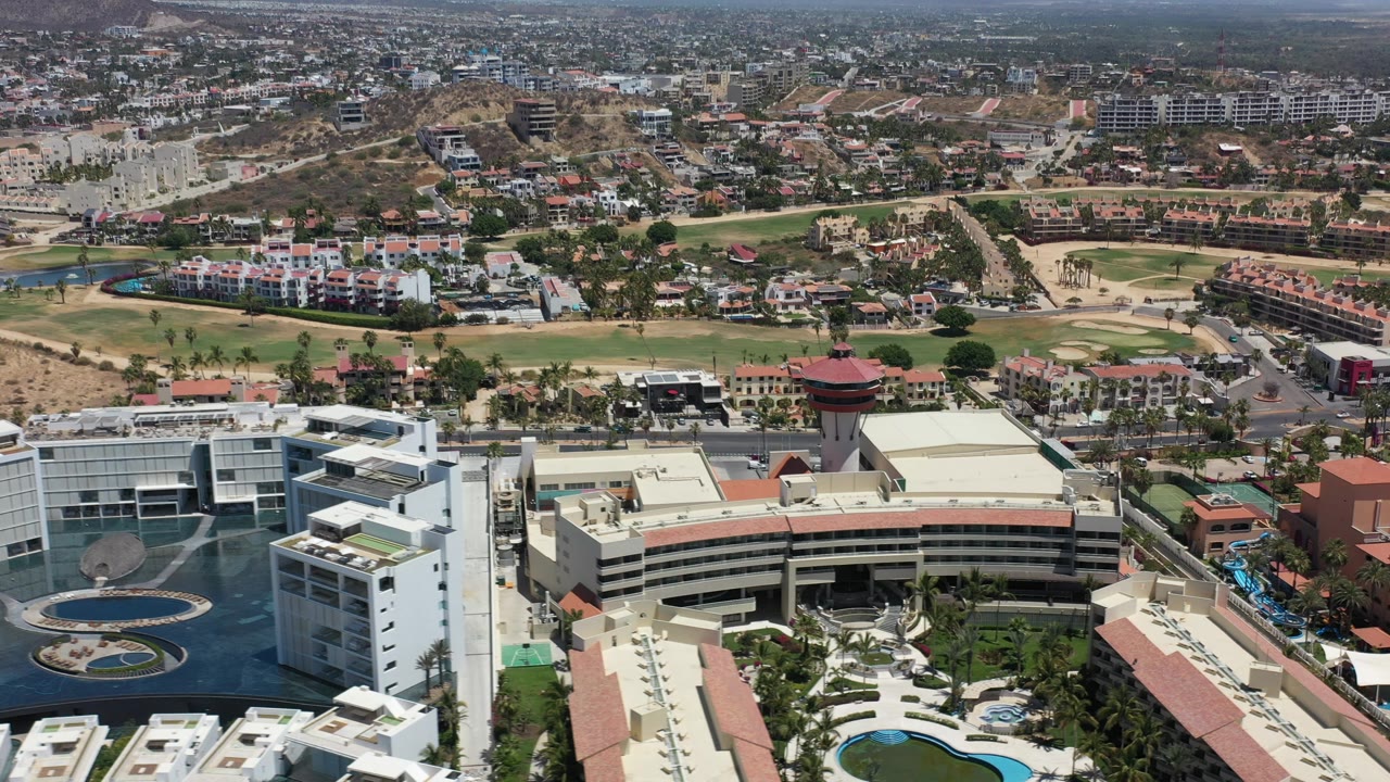 A wide aerial frame over Los Cabos, Mexico shows a large white building complex with pools in the foreground, roads and homes behind it, and dry hills beyond.