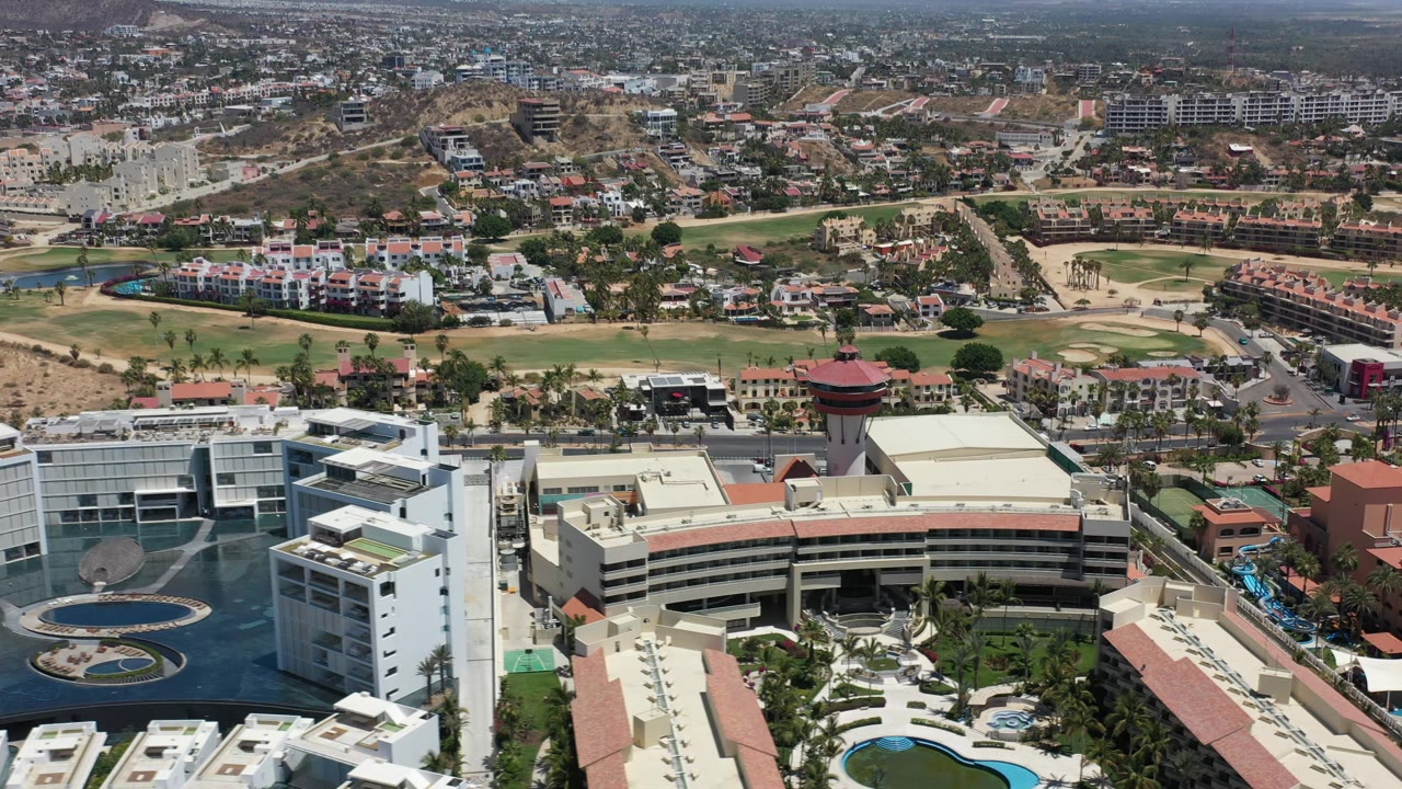 The aerial view over Los Cabos, Mexico centers more on a round-roofed tower and adjacent modern buildings, with roads, housing, and dry terrain across the background.