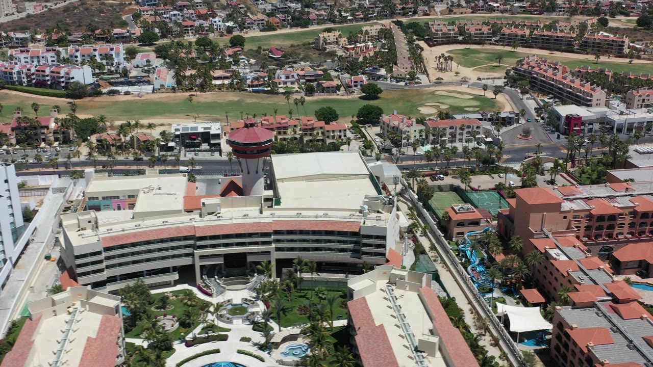 Opening aerial frame over Los Cabos, Mexico, with a curved multi-story building, red-roofed structures, palms, roads, and distant homes, with no visible people.