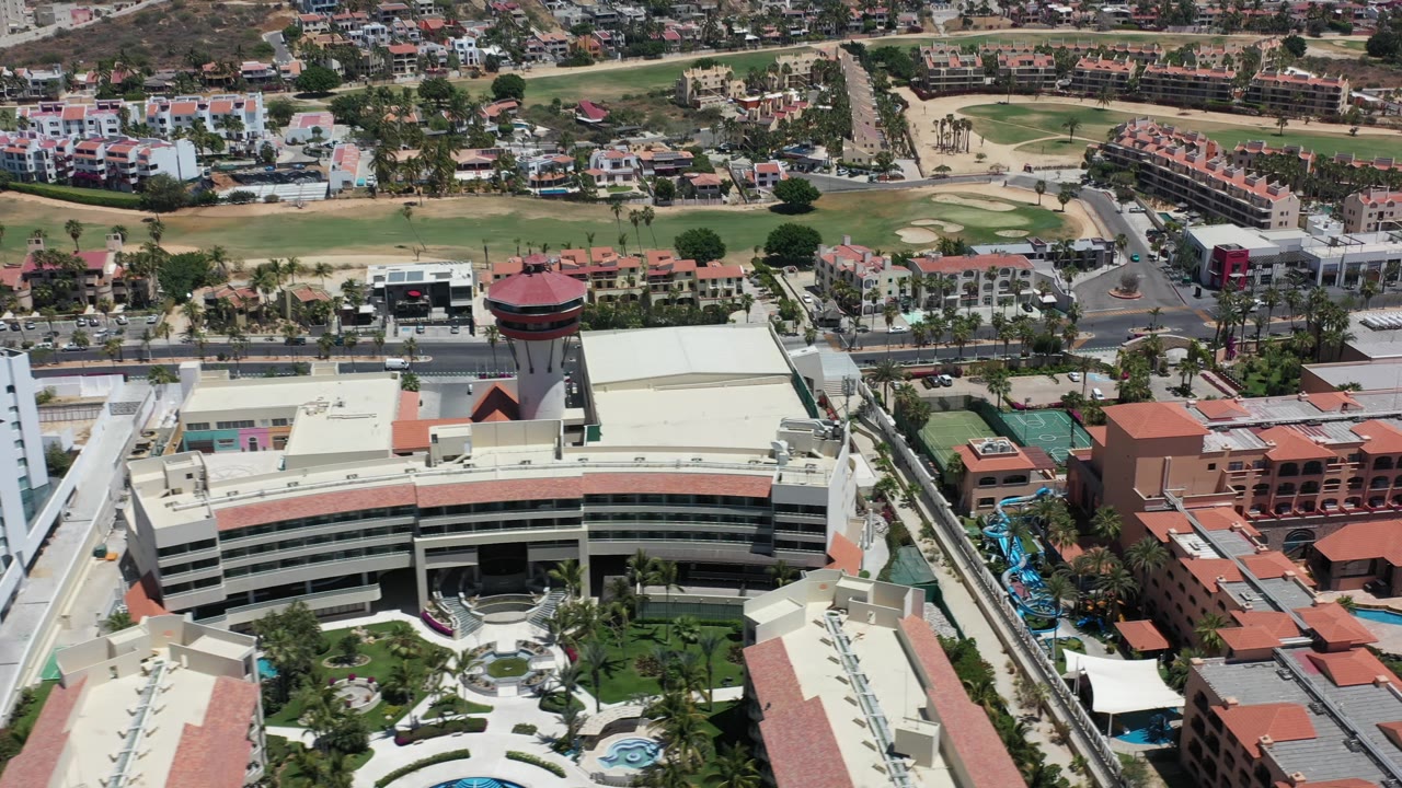 Los Cabos aerial frame with the curved building centered lower in view, a round tower to the right, red-roofed buildings, palms, roads, and a green course area in the background, with no visible people.