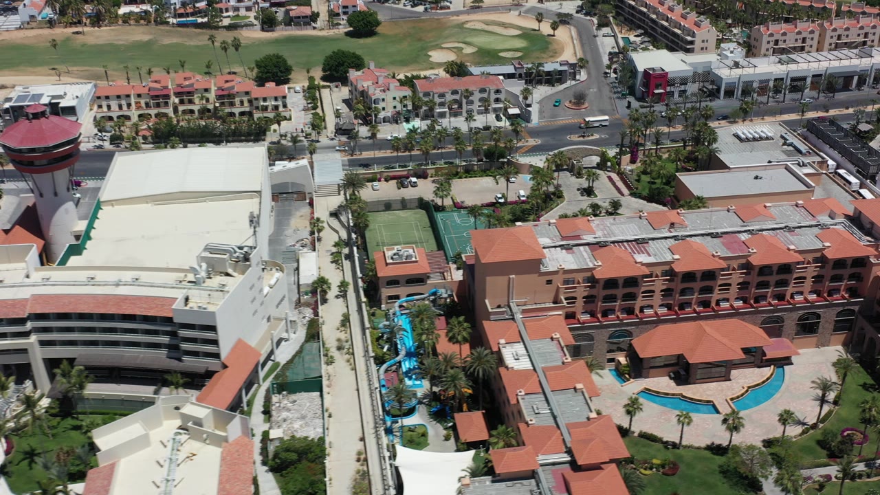 Opening aerial frame over Los Cabos resort buildings with red tile roofs, blue pools, roads, cars, tennis courts, golf-course grass, and no visible people.
