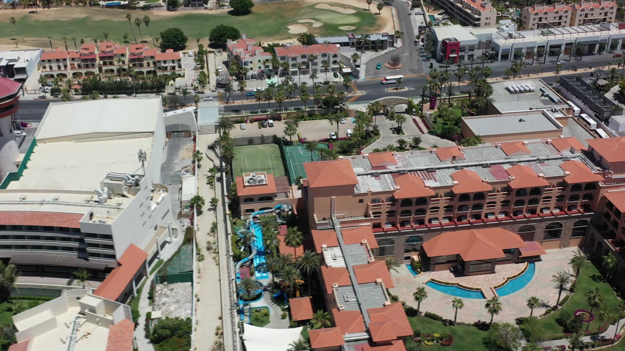 Aerial resort view with pools, hotel rooftops, white buildings, and roads.