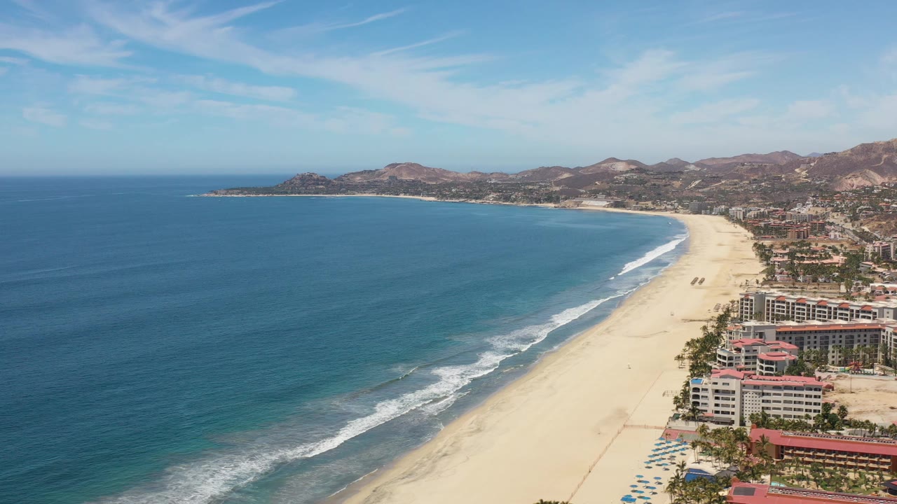 Wide aerial frame over Los Cabos, Mexico, showing a blue bay, pale sand beach, beachfront buildings at lower right, distant hills, and no visible people.