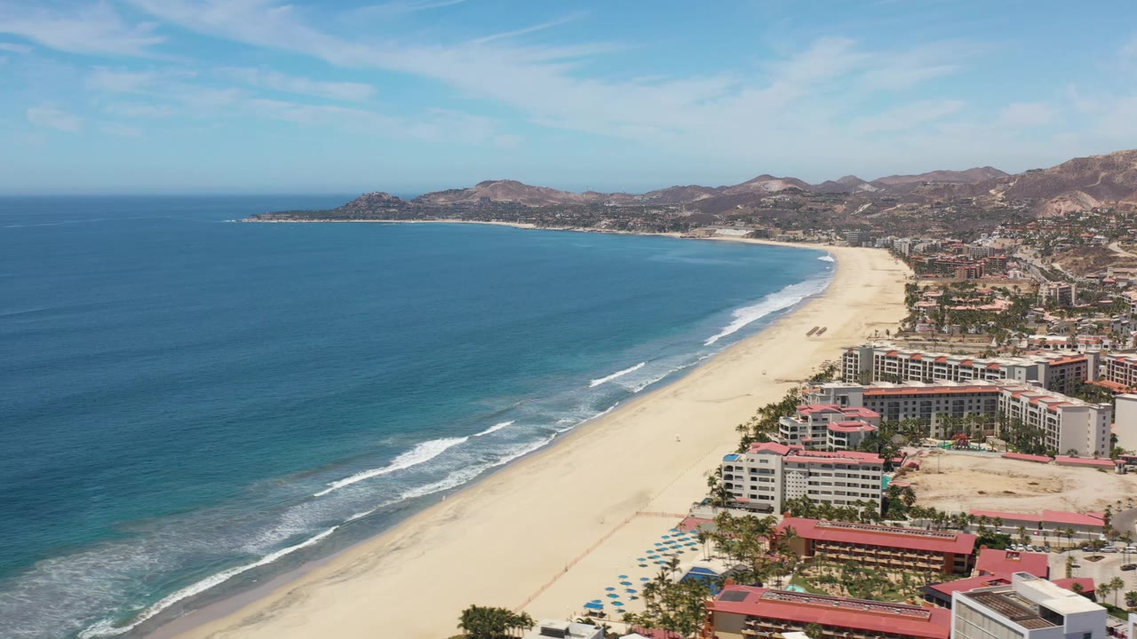 Wide aerial frame from Los Cabos, Mexico with the shoreline sweeping from the foreground into the distance, small surf bands along the sand, clustered beachfront buildings, and no visible people.