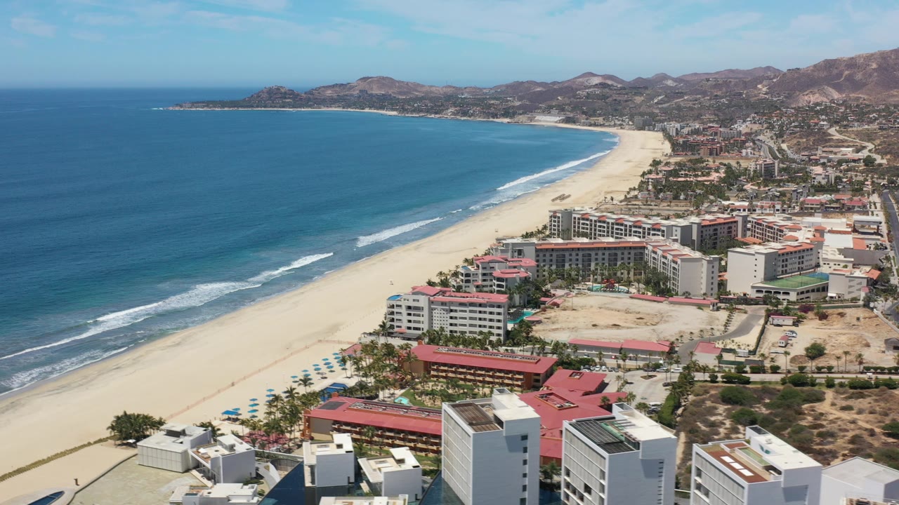 Opening aerial frame over Los Cabos, Mexico, looking along blue water, pale sand, beachfront hotel blocks, and dry hills, with no visible people.