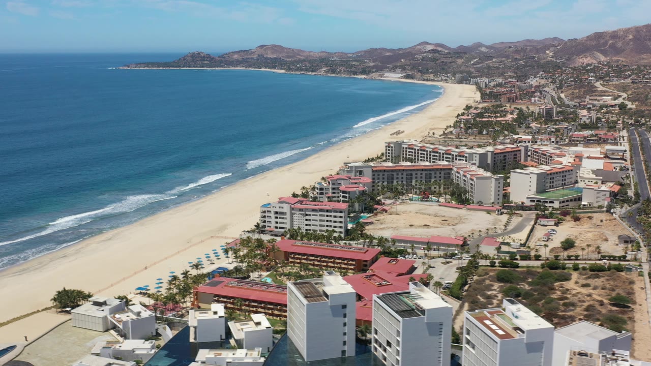 Drone-height coastal view with beach, hotels, road edge, and mountains beyond.
