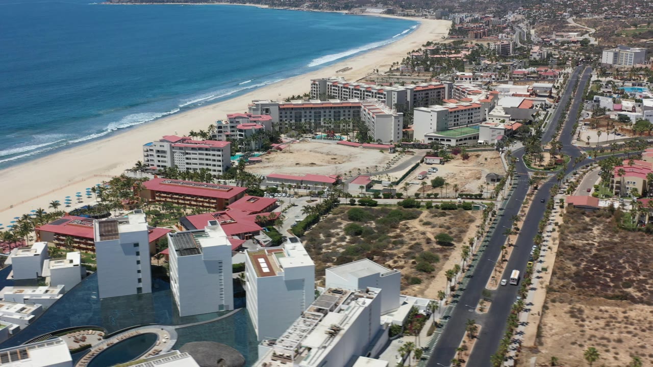 Drone view in Los Cabos showing resort buildings near the beach, a long road lined with cars, dry open lots, ocean surf, and no visible people.