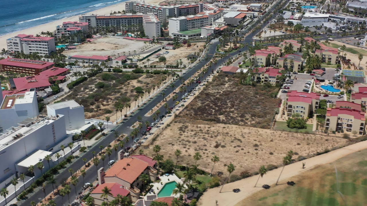 High aerial view over Los Cabos, Mexico with ocean surf at upper left, resort buildings, a straight road, red-roofed homes, pools, sandy lots, and no visible people.