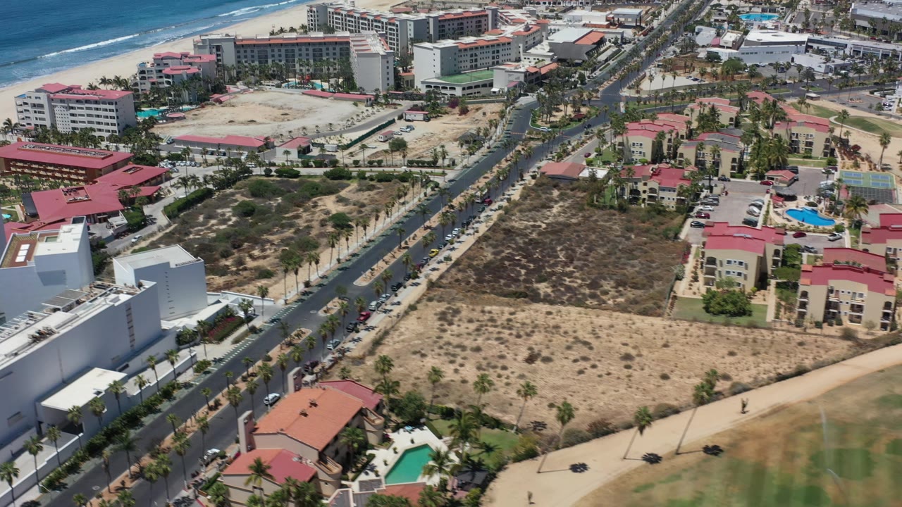 Aerial frame in Los Cabos, Mexico showing the shoreline beyond resort blocks, a palm-lined road through the center, red roofs and pools to the right, and no visible people.