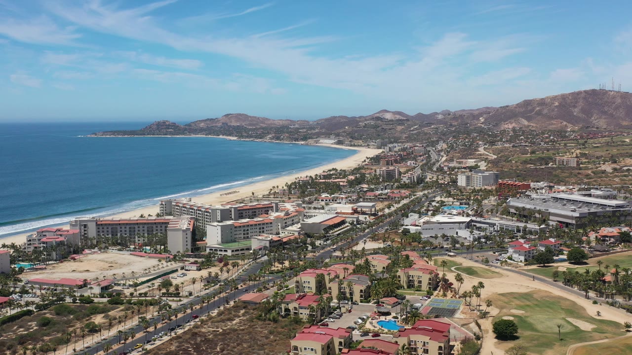 Wide aerial view of ocean, beach, city buildings, roads, and dry hills.