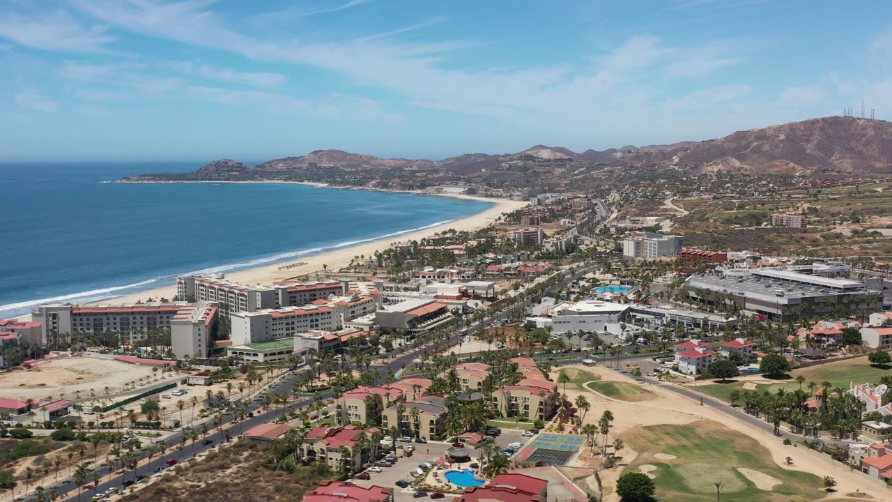 Aerial view over Los Cabos, Mexico, with a blue bay and pale beach at left, town buildings and palm-lined streets in the center, dry mountains in the background, and no visible people.