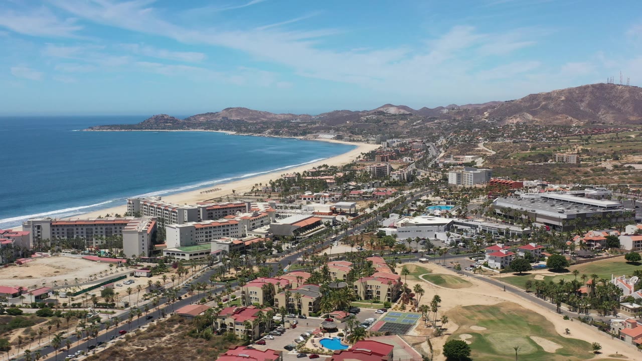 Wide aerial frame in Los Cabos, Mexico, showing the curved shoreline, blue water, light sand, coastal buildings, roads, palm trees, arid hills, and no visible people.