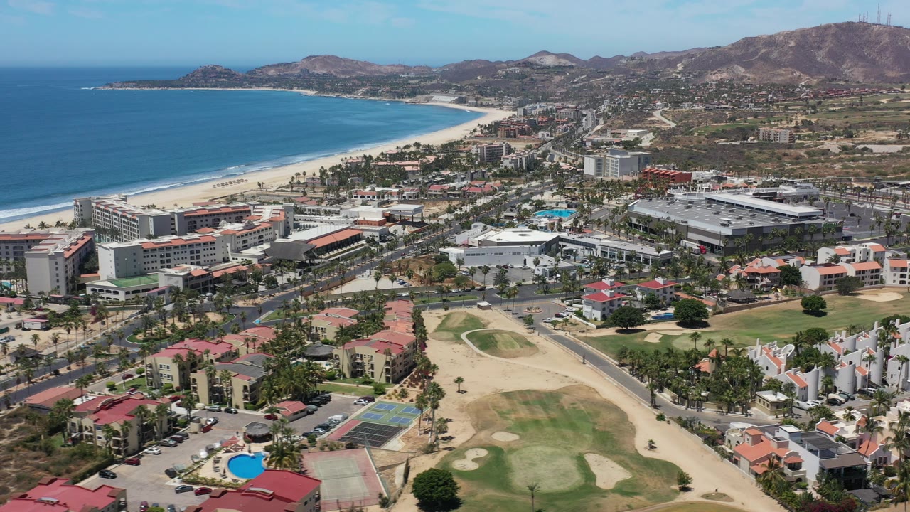 Aerial frame over Los Cabos, Mexico, showing a curved blue coastline, beachfront buildings, foreground golf course, condominium clusters, roads, dry hills, and no visible people.