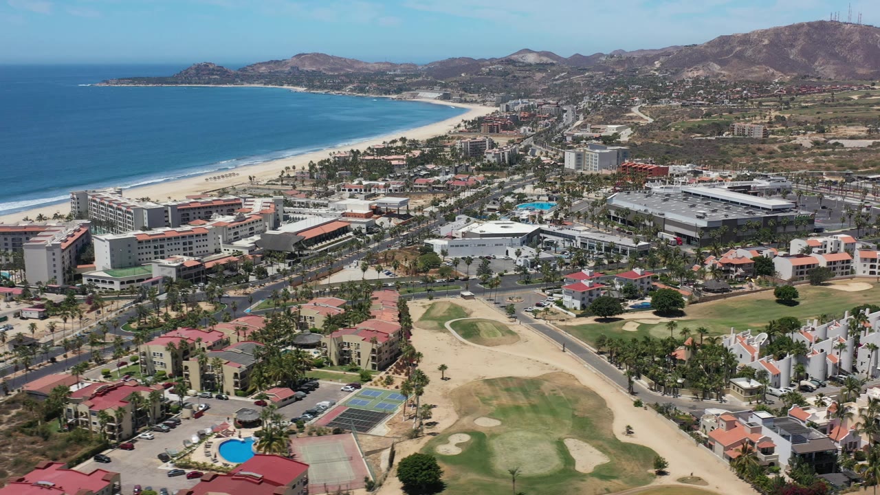 Los Cabos, Mexico aerial view with the beach and ocean on the left, dense condominium and resort-style buildings in the center, golf greens below, hills behind, and no visible people.