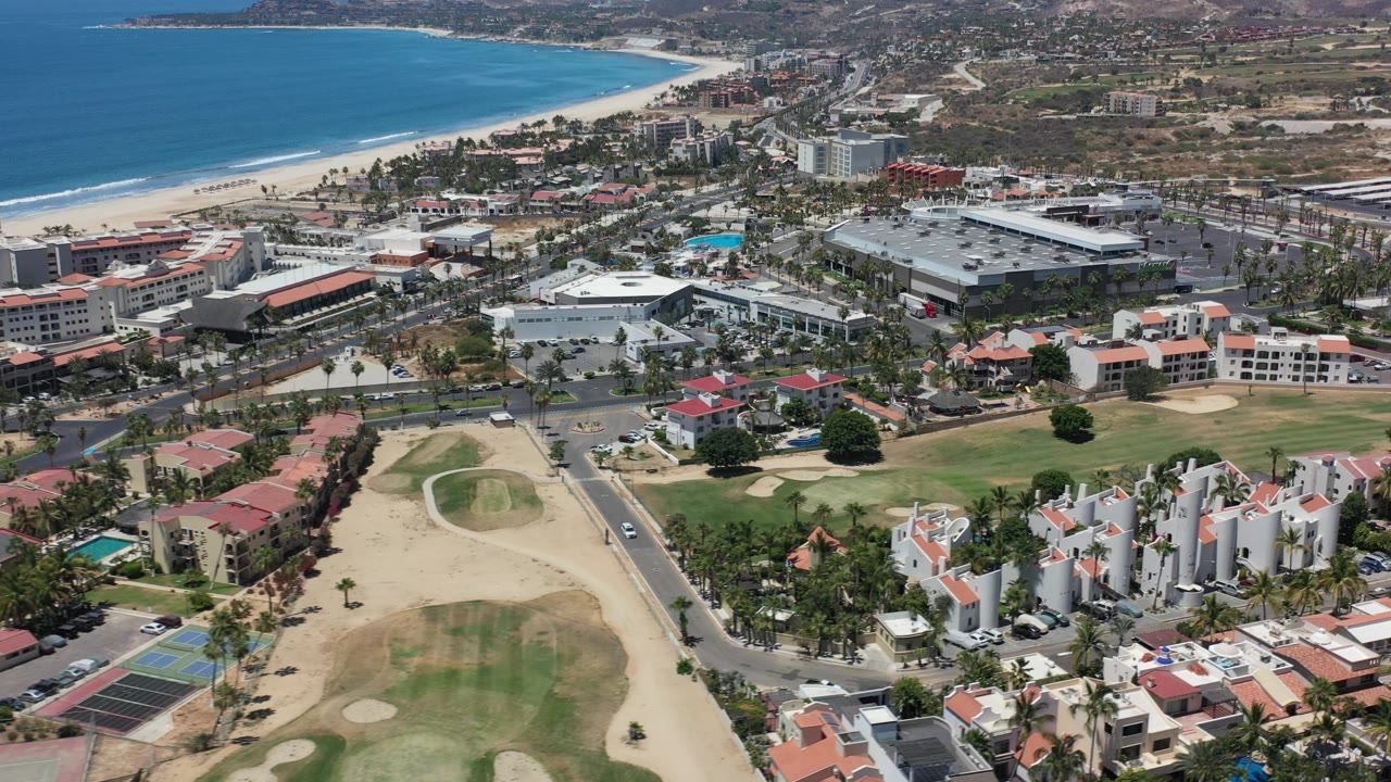 High aerial frame over Los Cabos, Mexico with blue ocean, sandy beach, low-rise buildings, palm-lined roads, golf course grass, private homes, and no visible people.