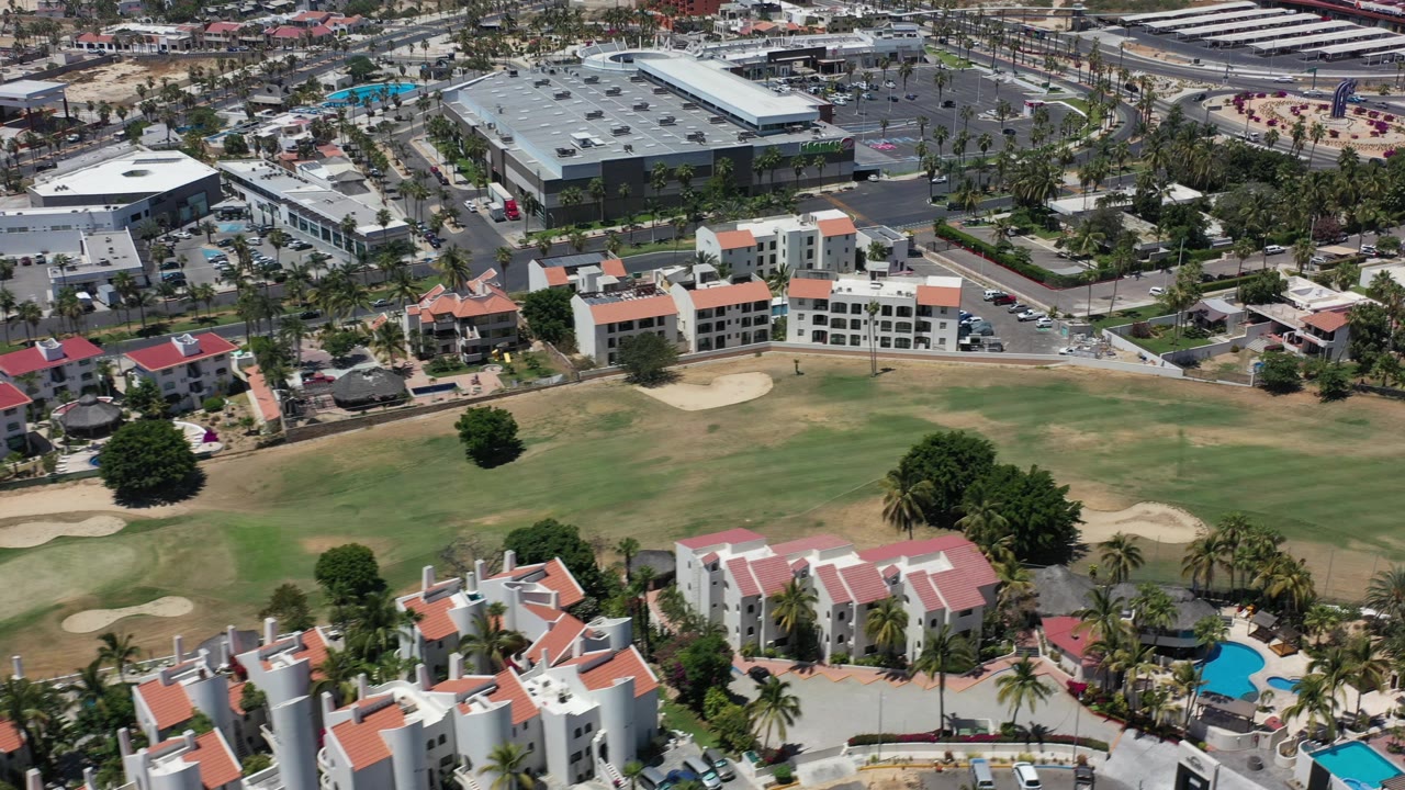 Opening aerial frame over Los Cabos, Mexico, with a golf fairway, sand bunkers, white resort buildings, red roofs, blue pools, roads, palm trees, and no visible people.