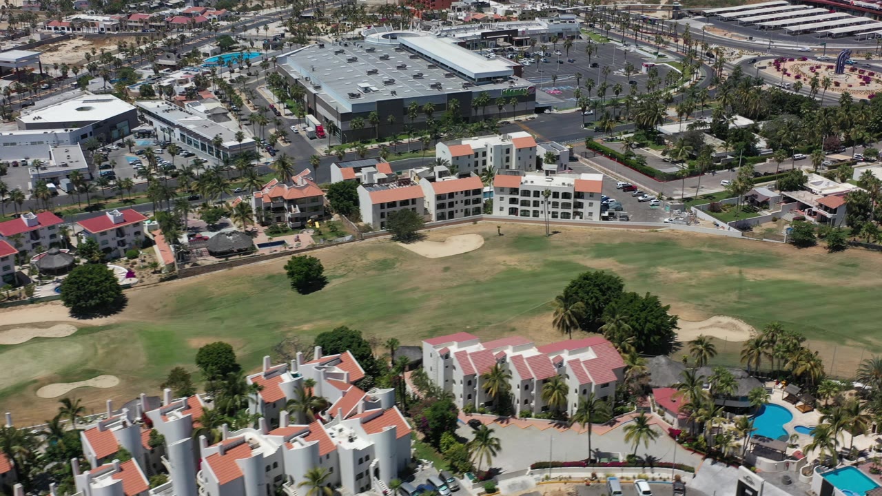 Aerial frame in Los Cabos, Mexico, looking across tan and green golf-course grass toward low-rise buildings, roads, parking lots, palm trees, and no visible people.