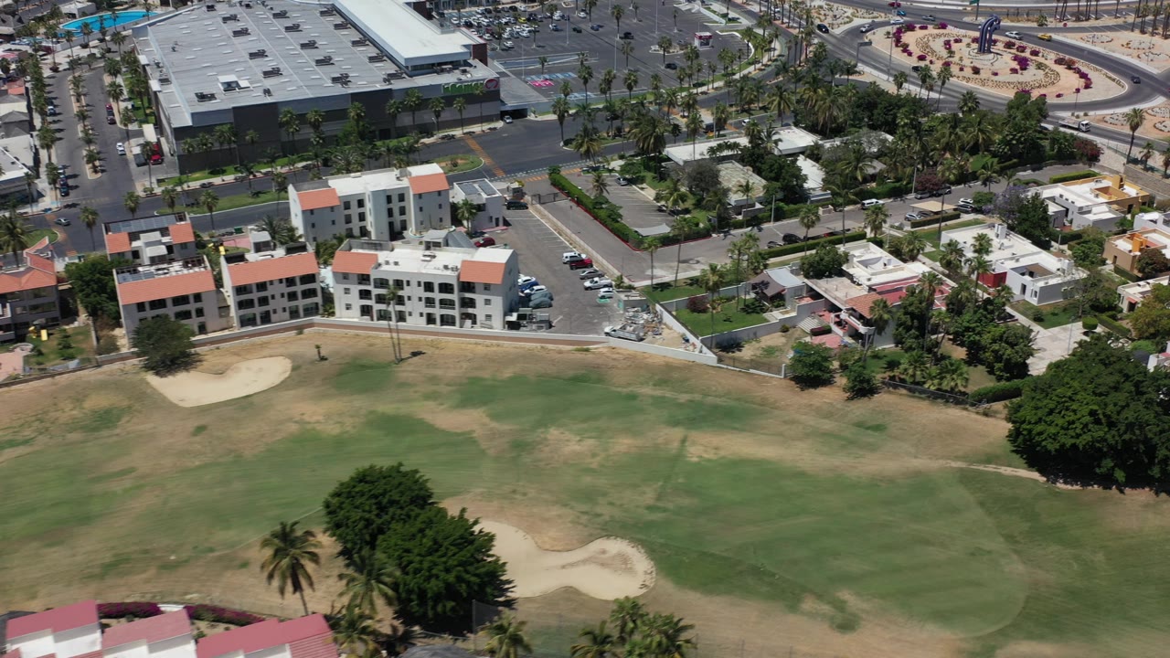 Aerial frame from Los Cabos, Mexico showing a golf fairway with tan dry patches, sand bunkers, low-rise buildings, parked cars, roads, palms, and no visible people.