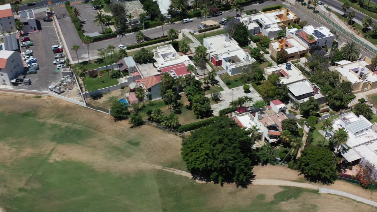 A high aerial frame over Los Cabos, Mexico shows flat-roof homes, a dry green-brown field, dense trees, parked cars, and no visible people.