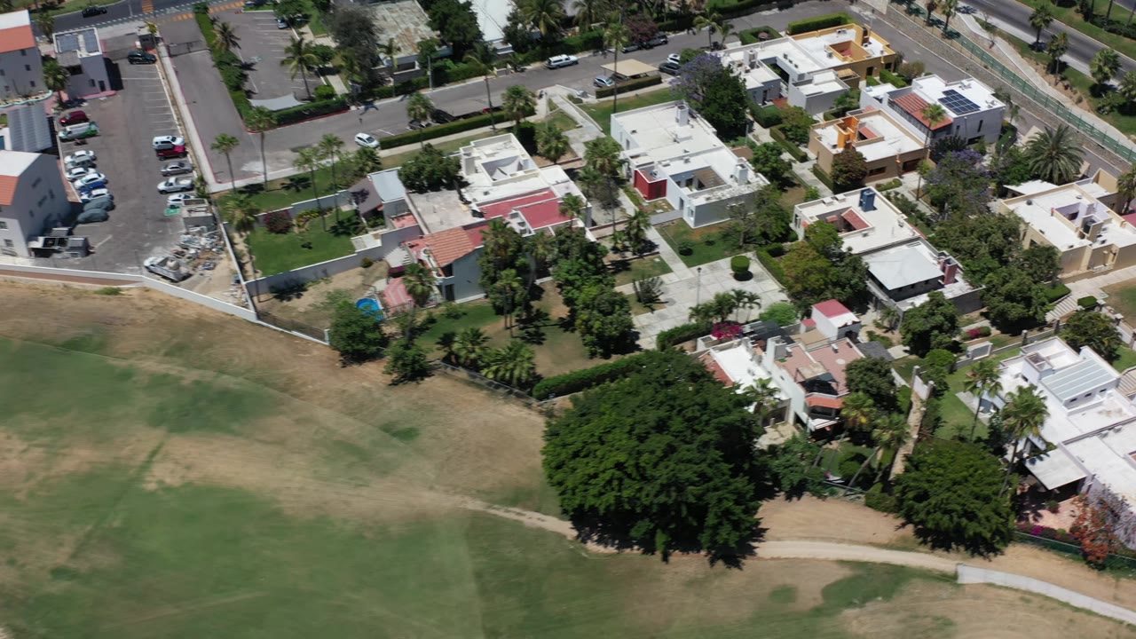 The Los Cabos aerial view continues over homes and garden walls, with a large tree canopy at the field edge and no visible people.
