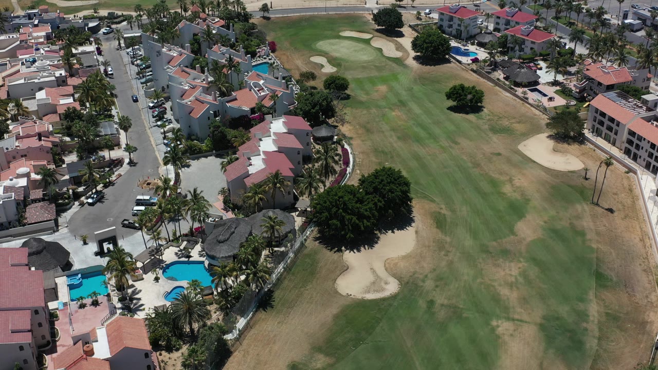Opening aerial frame over Los Cabos condominiums, blue pools, palm-lined roads, and a golf fairway with sand bunkers, with no visible people.