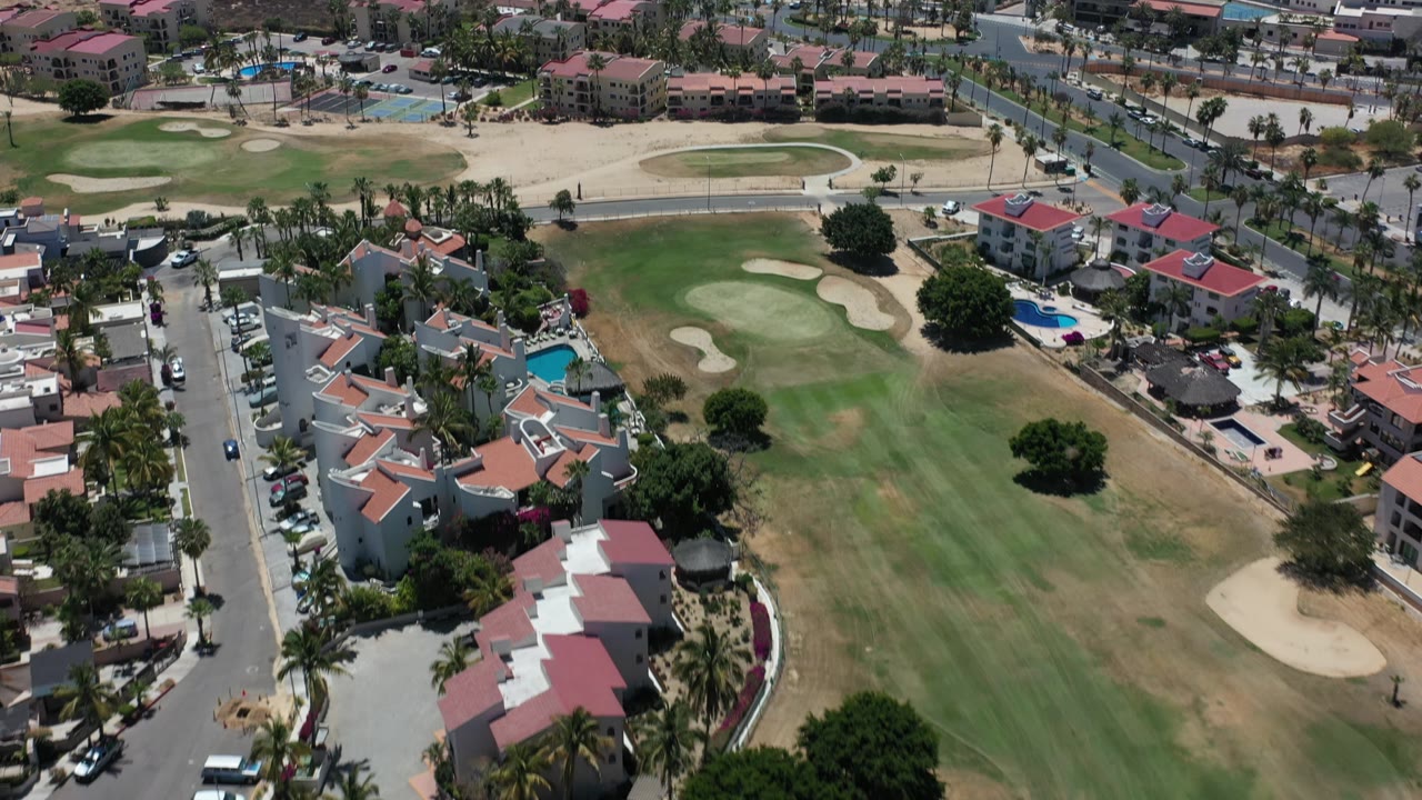 Elevated Los Cabos frame with fairways and sand bunkers in the center, villas and parked cars below, beachfront buildings above, and no visible people.