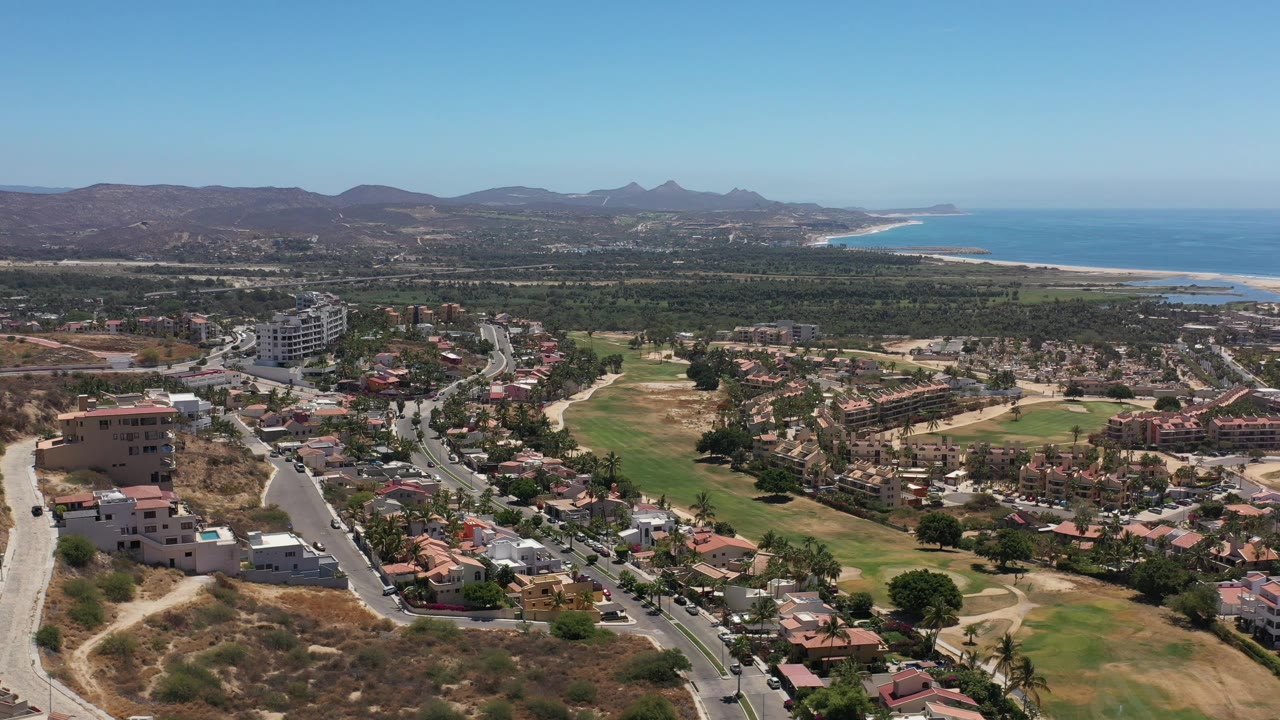 Wide Los Cabos aerial frame with residential streets in front, green fairway-like areas in the middle, mountains on the horizon, ocean to the right, and no visible people.