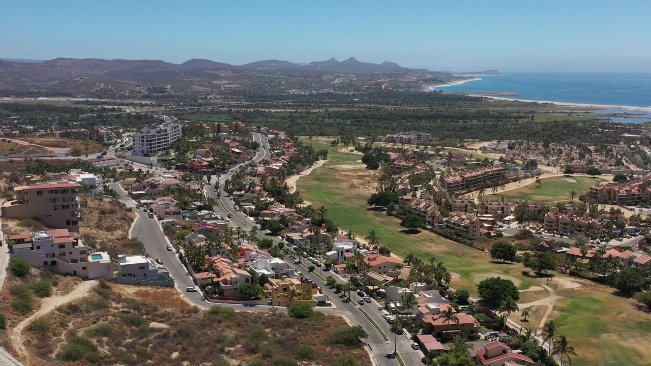 Aerial view of residential roads, homes, fairway grass, hills, and distant coastline.