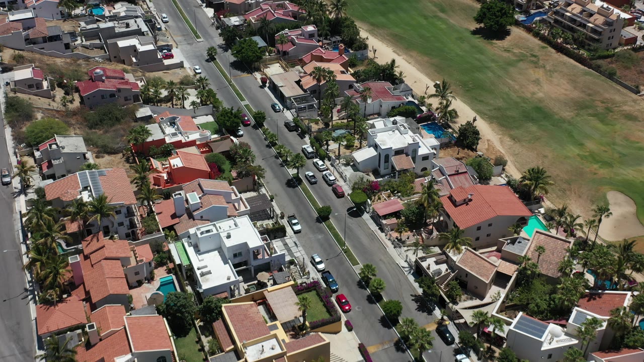 Top-view aerial over Los Cabos, Mexico, with the road slightly more centered, compact homes and courtyards at left, a white modern house below, and dry open land to the right, with no visible people.